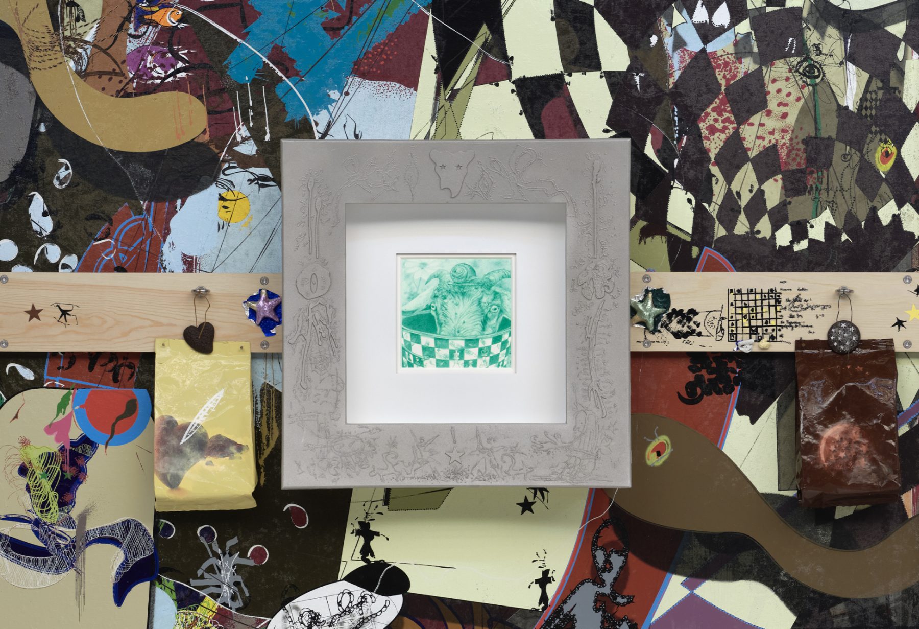 An overhead shot of a collage with cutouts of brown snakes, checkered patterns, and a wooden beam cutting across horizontally. A framed sketch in green ink rests in the center.