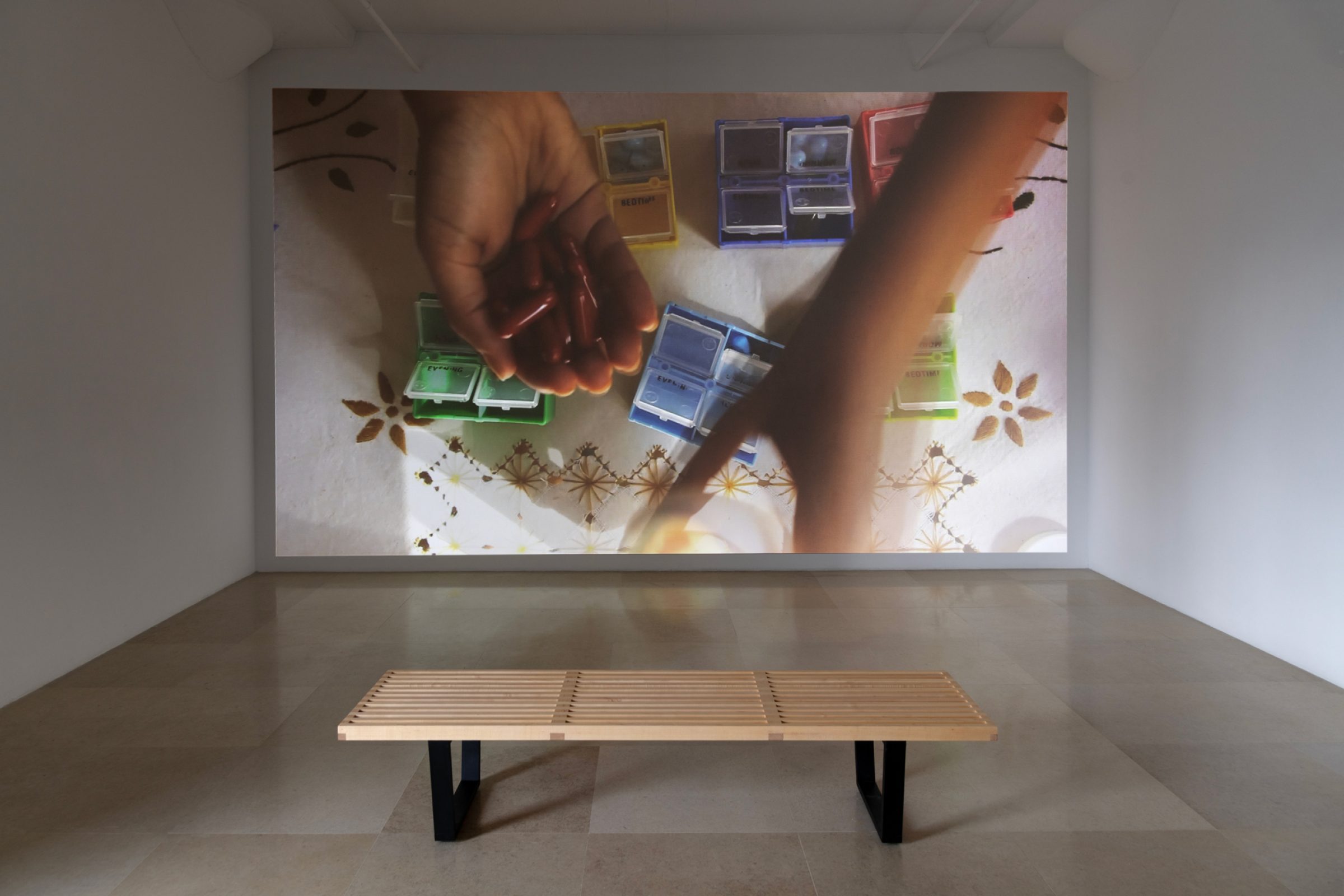 Still of a projected video in a white walled room with a bench. The still depicts a hand holding bright red pills on top of a table with a floral patterned table cloth and colorful pill organizers.