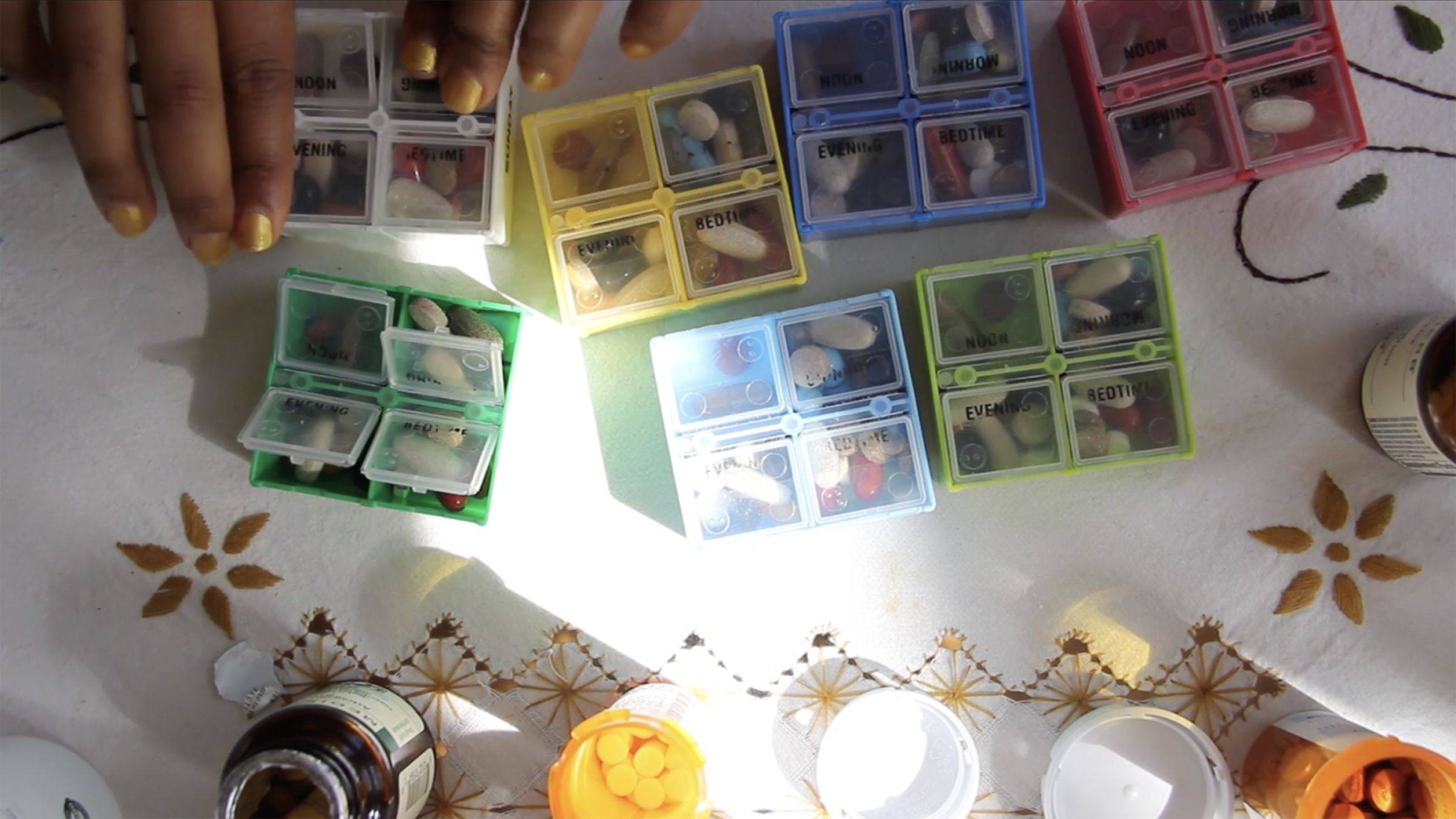 Overhead view of two hands sorting pills into seven pillbox compartments on an embroidered tablecloth. The compartments are yellow, blue, pink, green, and white, and the pills inside are various colors. A patch of bright sunlight falls across the cloth, and open pill bottles sit along the edge of the frame.