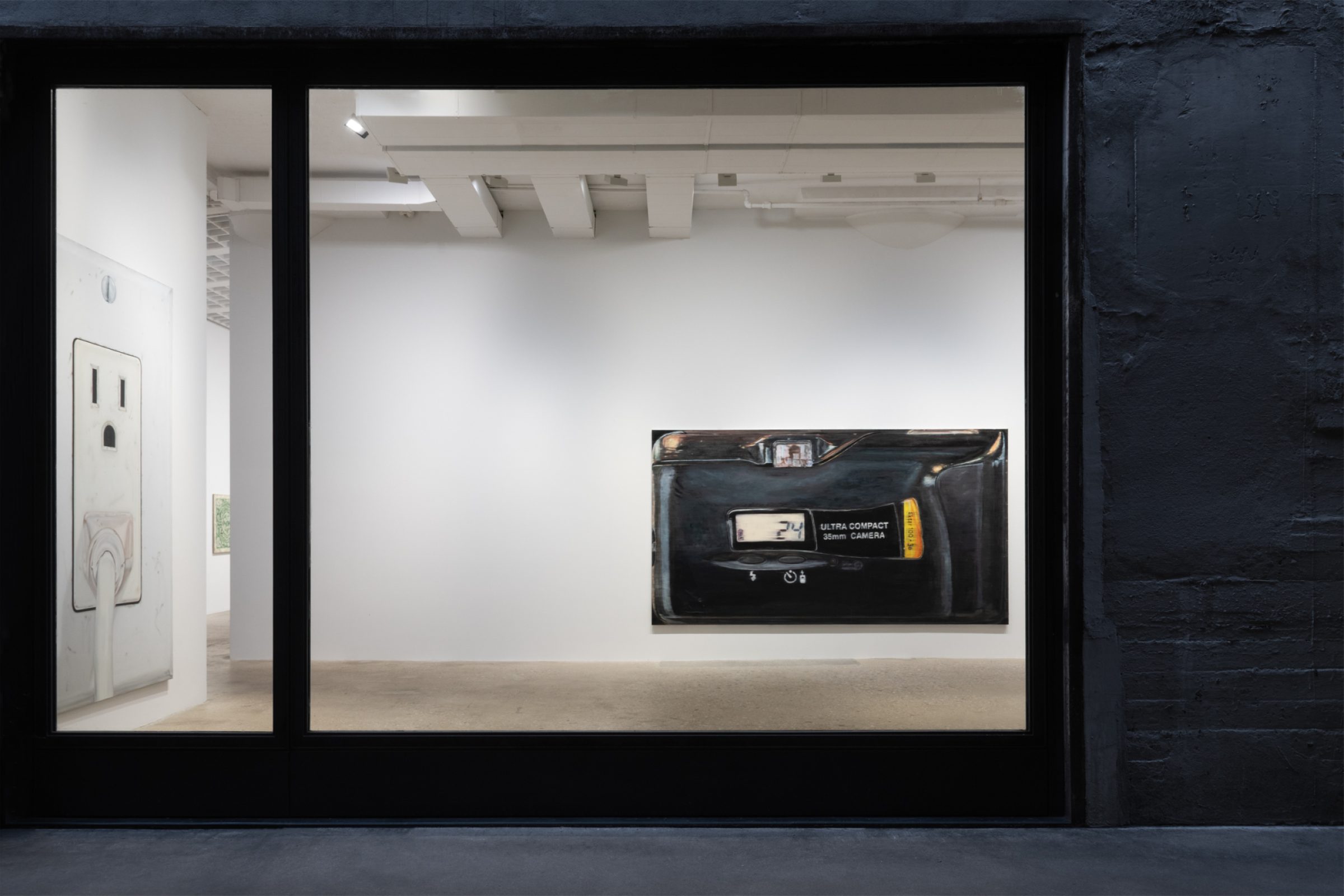 Photographed through the front window of a gallery, a painting of a a painting of the viewfinder side of an Ultra Compact 35mm Film Camera faces out; on the left wall one can see a painting of an American wall outlet with a plug plugged into the bottom socket.