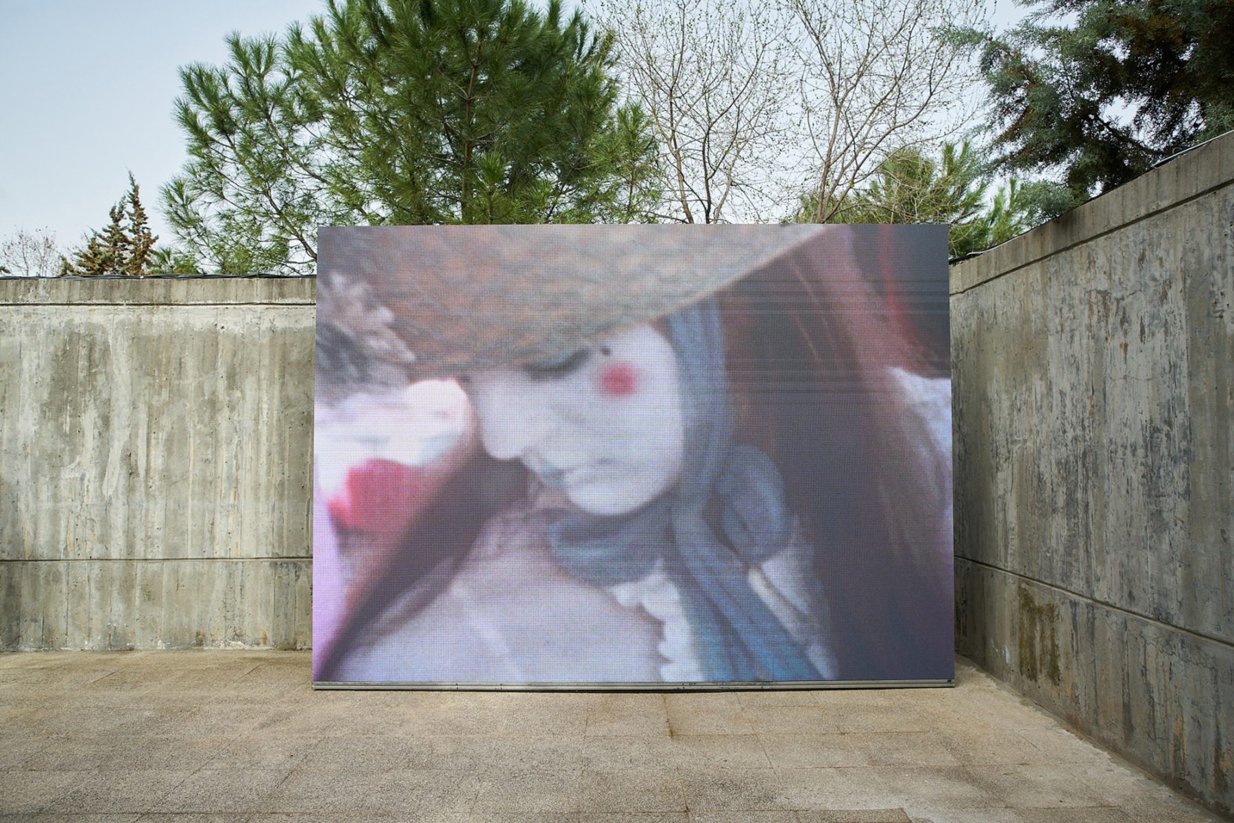 Installation shot of an image of a woman with white facepaint and a hat tied under her chin with a blue ribbon stands outside against a concrete wall.