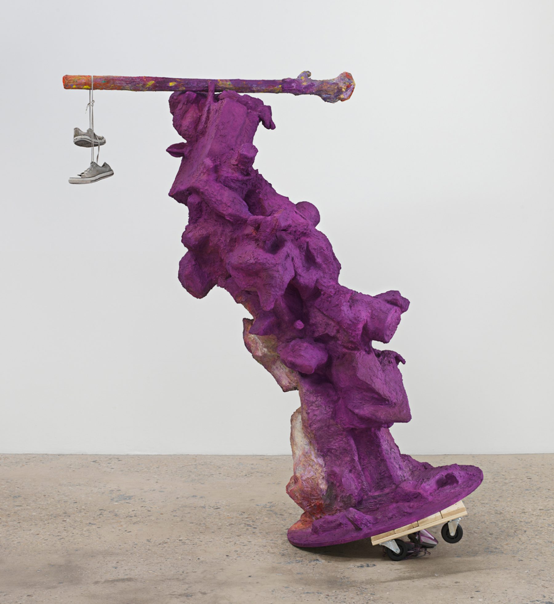 A room with white walls, a white column, and concrete floors with natural light streaming in from the ceiling. A curved purple statue stands in the center with a pair of sneakers hanging from the top.