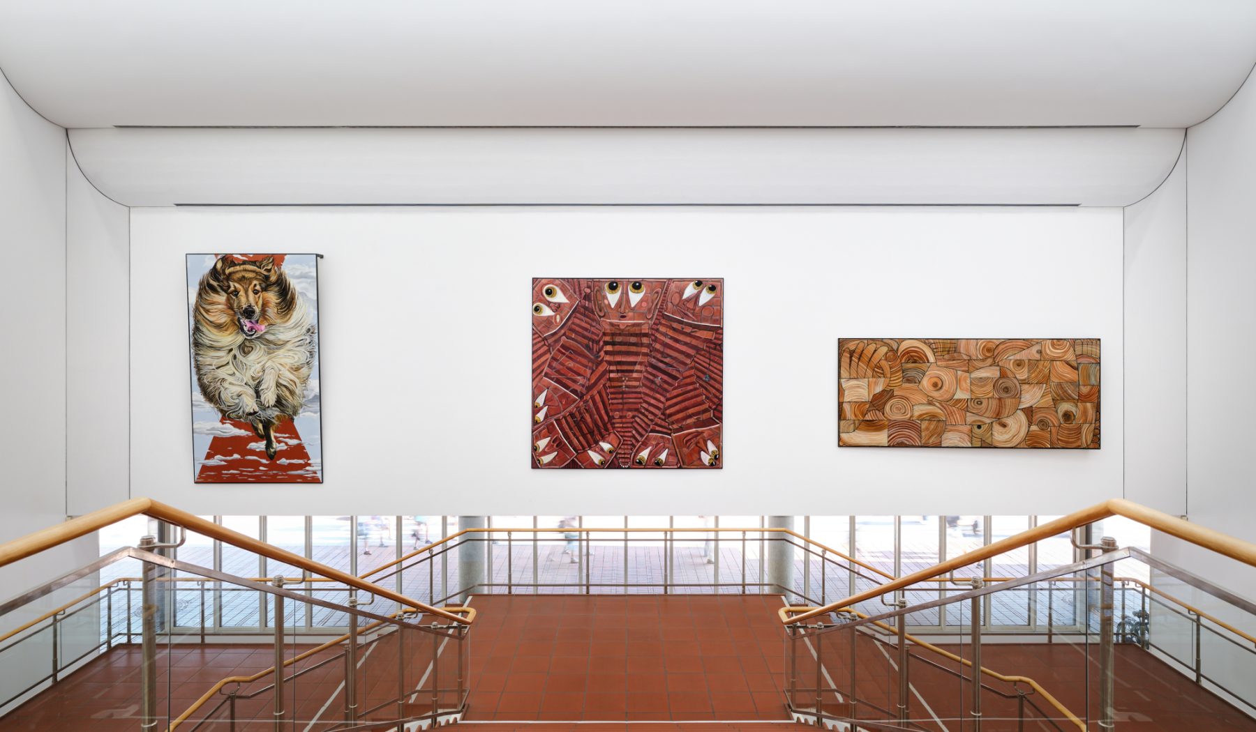 An installation view of a staircase with wood railing and glass panels leading down. On a white wall across from the staircase hangs three paintings: one is of a golden collie dog, one is of red staircases and eyes, and one is of brown swirls.