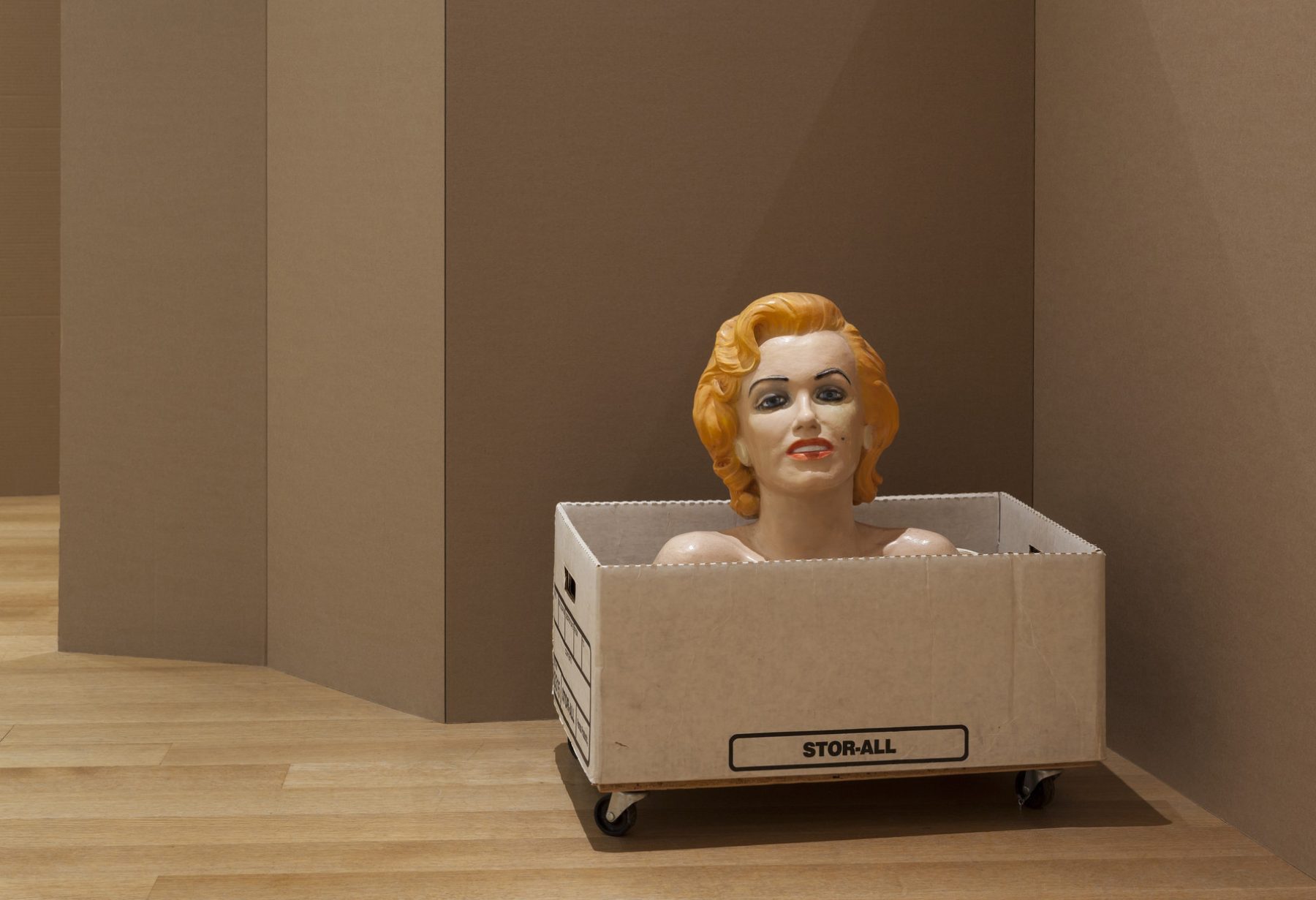 An image of a mannequin’s head popping out of a cardboard box in front of brown walls.