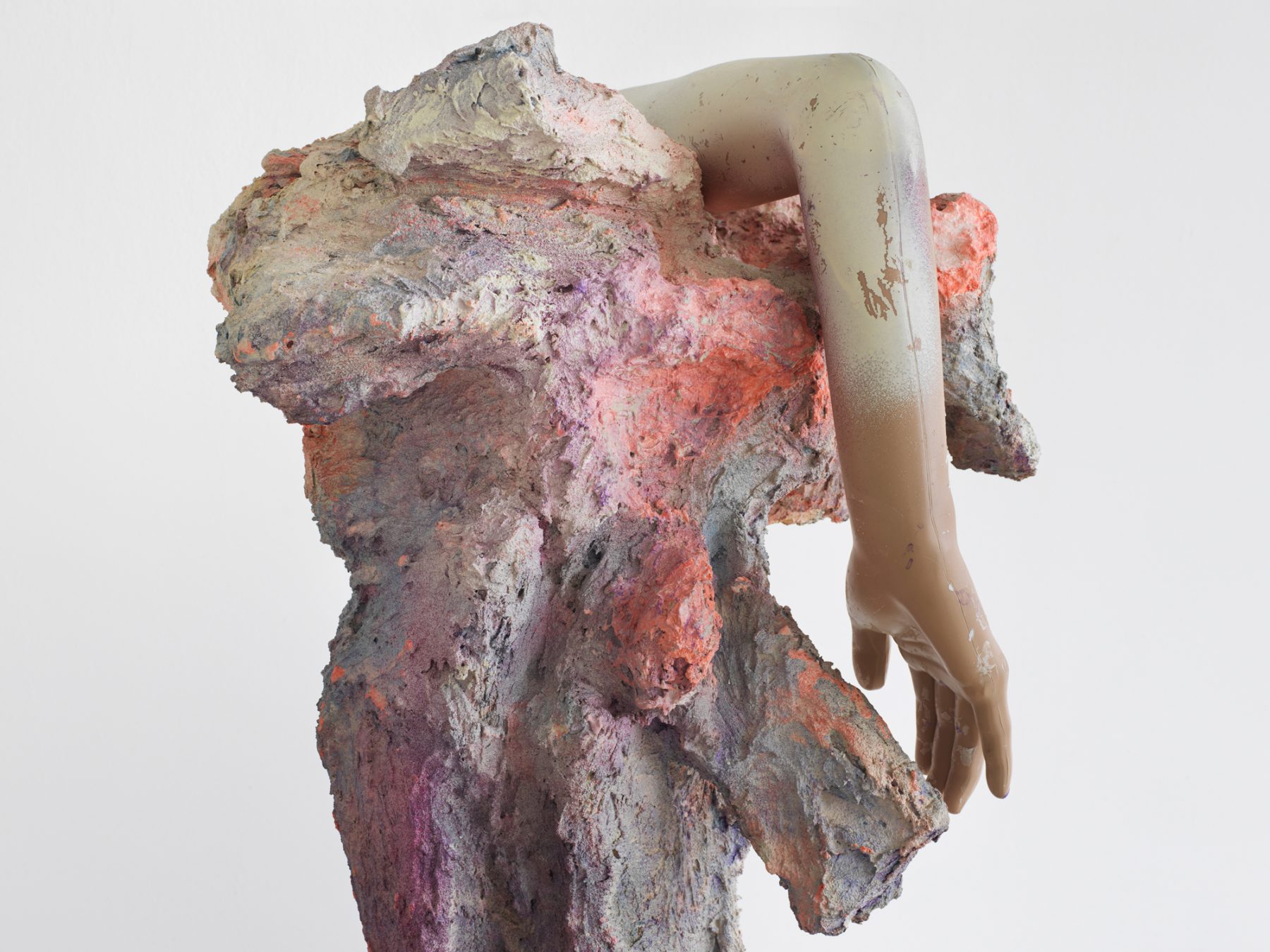 Close up of a textured statue covered in grey, purple, and peach paint, and a plastic human arm bent over the top of the statue.