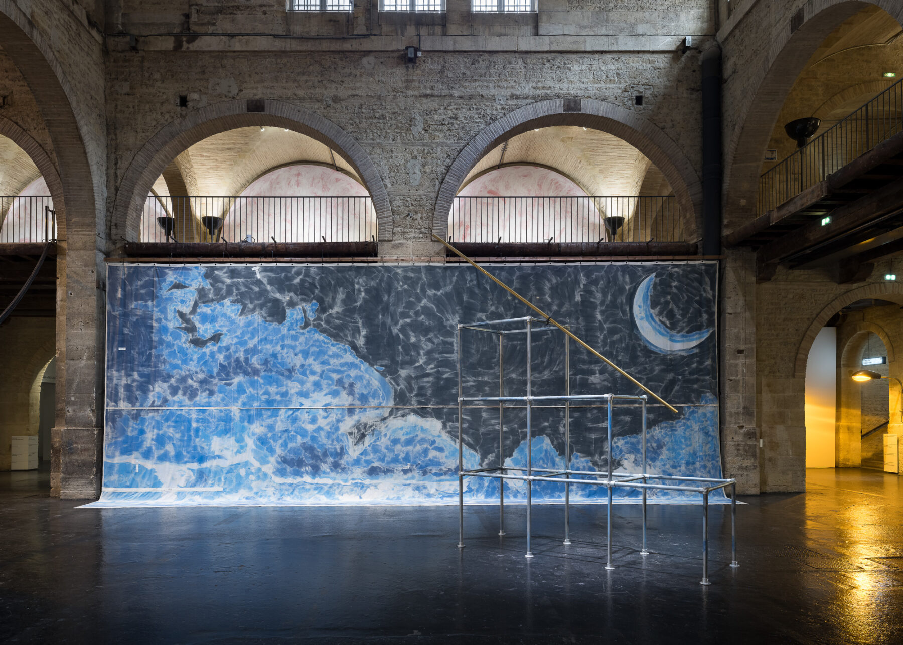 Interior of space with a large ocean mural on the wall and metal framework to the right side of the picture. The floors are black and the building is stone, with two stone arches directly above the mural.